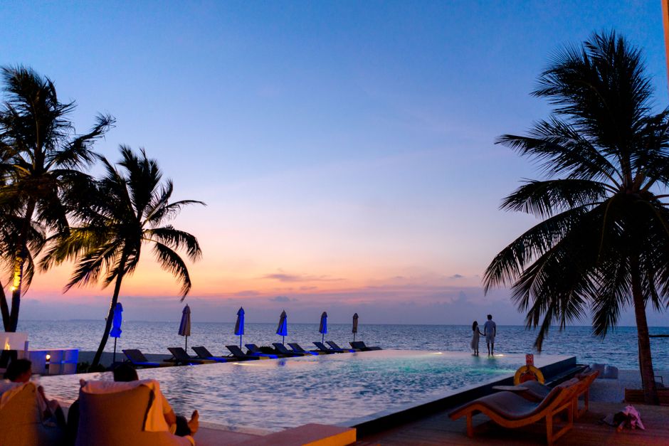 A couple enjoys a picturesque sunset by the seaside infinity pool, surrounded by palm trees.